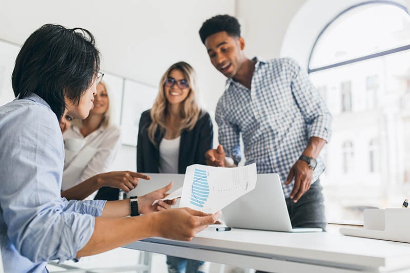 asian-manager-wristwatch-sitting-his-workplace-holding-paper-with-graph-indoor-portrait-african-programmer-checkered-shirt-talking-with-other-employees-office